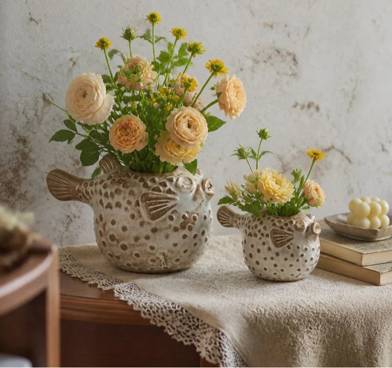 Two ceramic fish-shaped flower vases filled with peach roses and yellow flowers, displayed on a draped tablecloth against a textured stone wall.