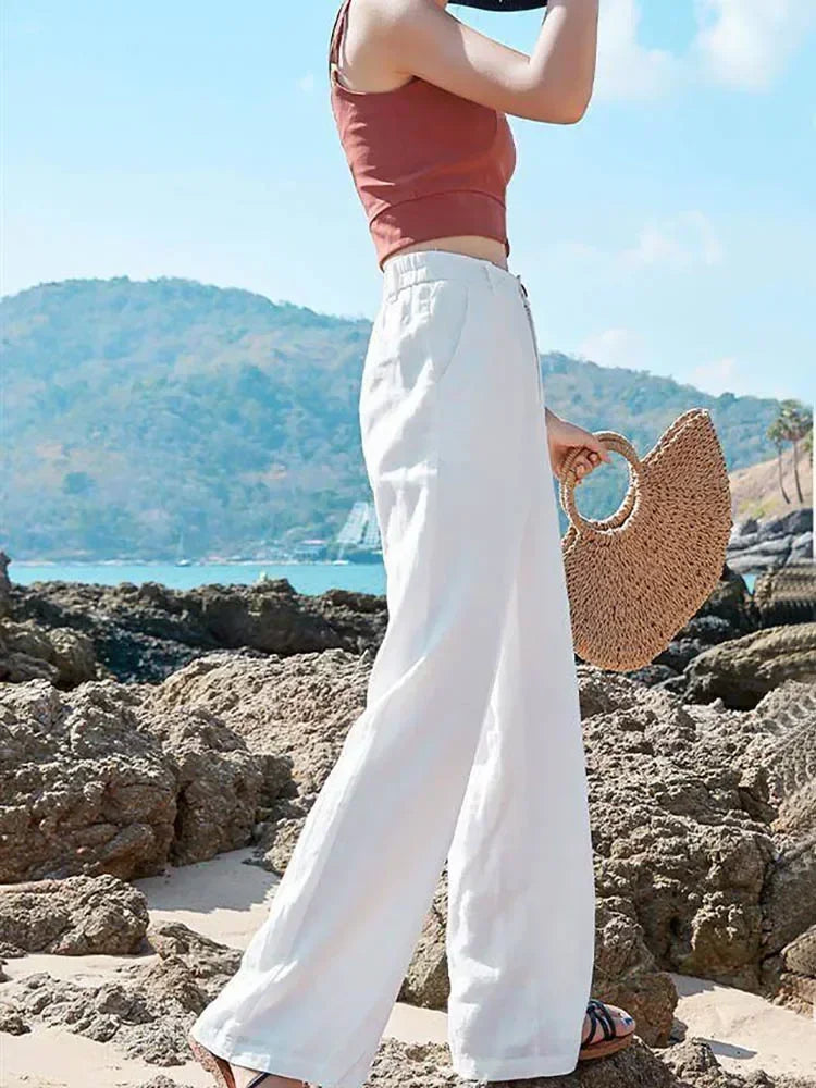 Person wearing a red top and white pants holding a straw bag on a rocky beach with mountains in the background.