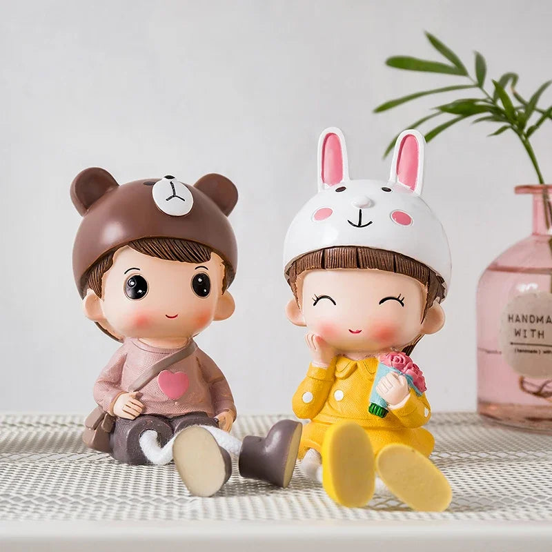 Two cartoon figurines, one resembling a boy with a bear hat and the other resembling a girl with a rabbit hat, sitting on a surface with a plant and bottle in the background.