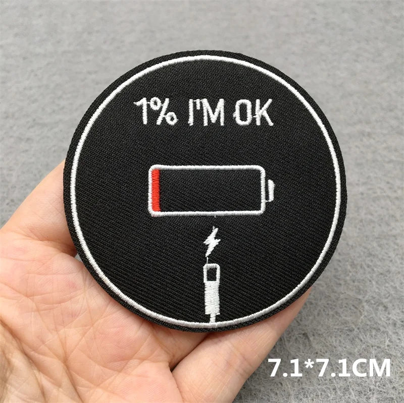 Circular patch with battery symbol and '1% I'm OK' text held in a hand on a gray background.
