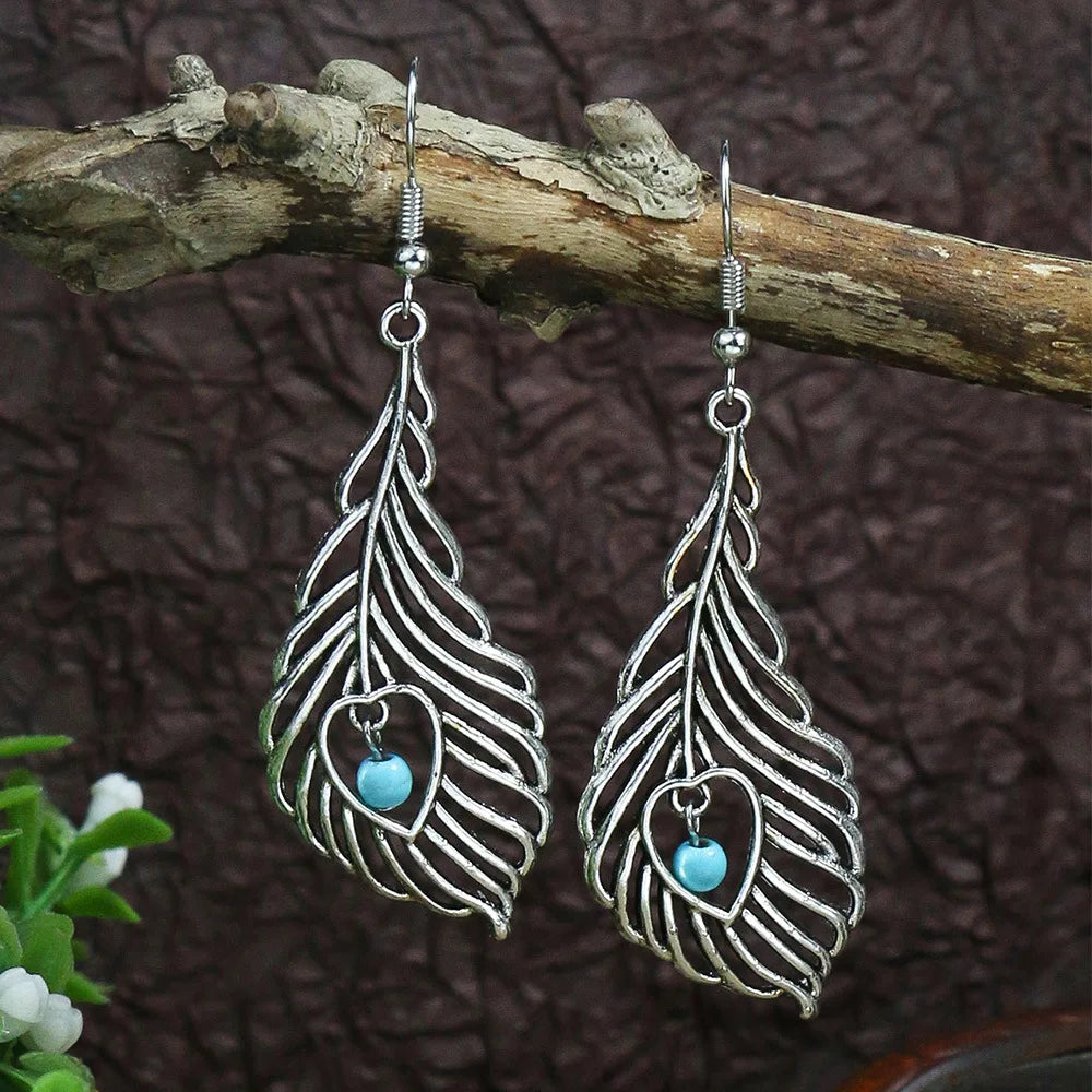 Silver feather-shaped earrings with turquoise beads on a wooden branch.