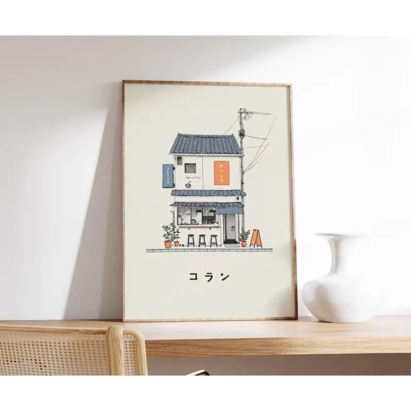 A framed art print featuring a hand-drawn illustration of a small Japanese-style cafe building, leaning against a white wall on a wooden shelf next to a white ceramic vase.