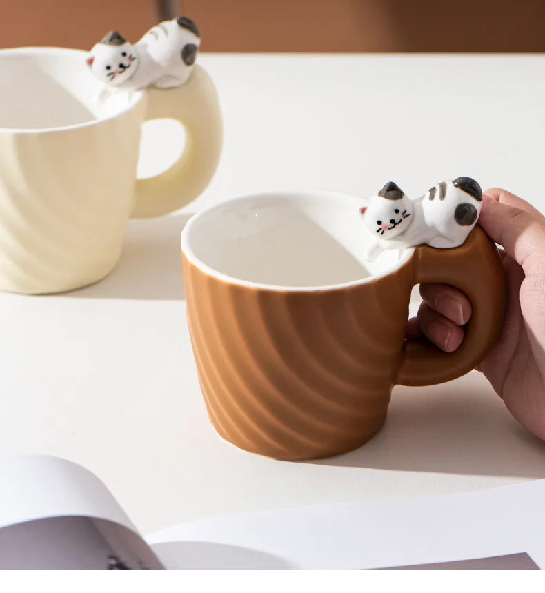 Brown ceramic mug with cat-shaped handle held by a hand, on a white surface.