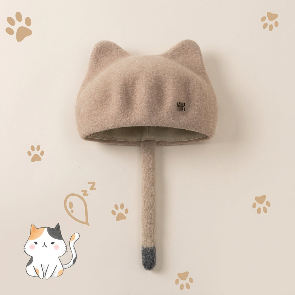 A beige cat-ear hat with a long tail hanging from the bottom, shown on a light beige background with paw-print graphics and a cartoon cat illustration. 