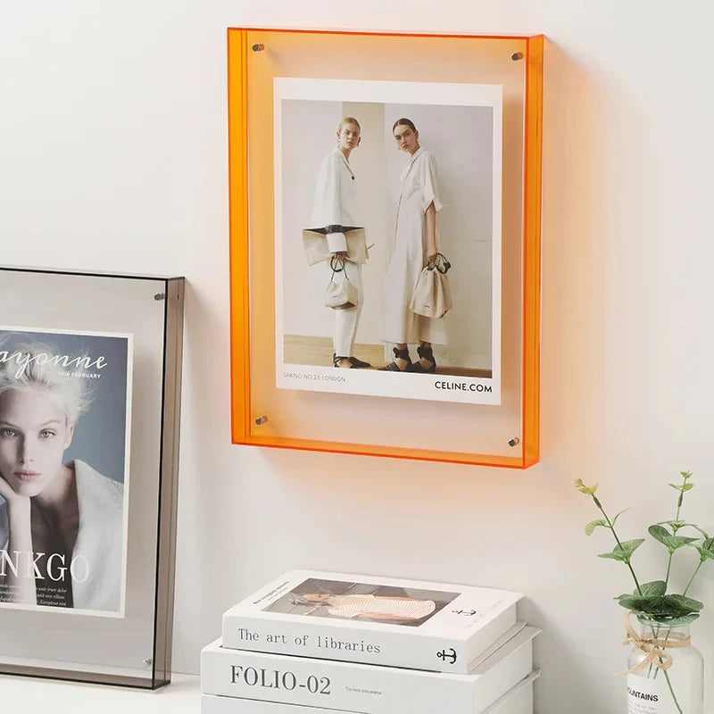 Transparent orange acrylic picture frame mounted on a white wall, displaying a fashion photograph above stacked books beside a small plant.