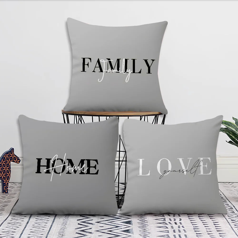 Three gray square throw pillows with black and white decorative text reading FAMILY together, HOME with script, and LOVE together, arranged on a black wire chair and a patterned rug against a white wall.