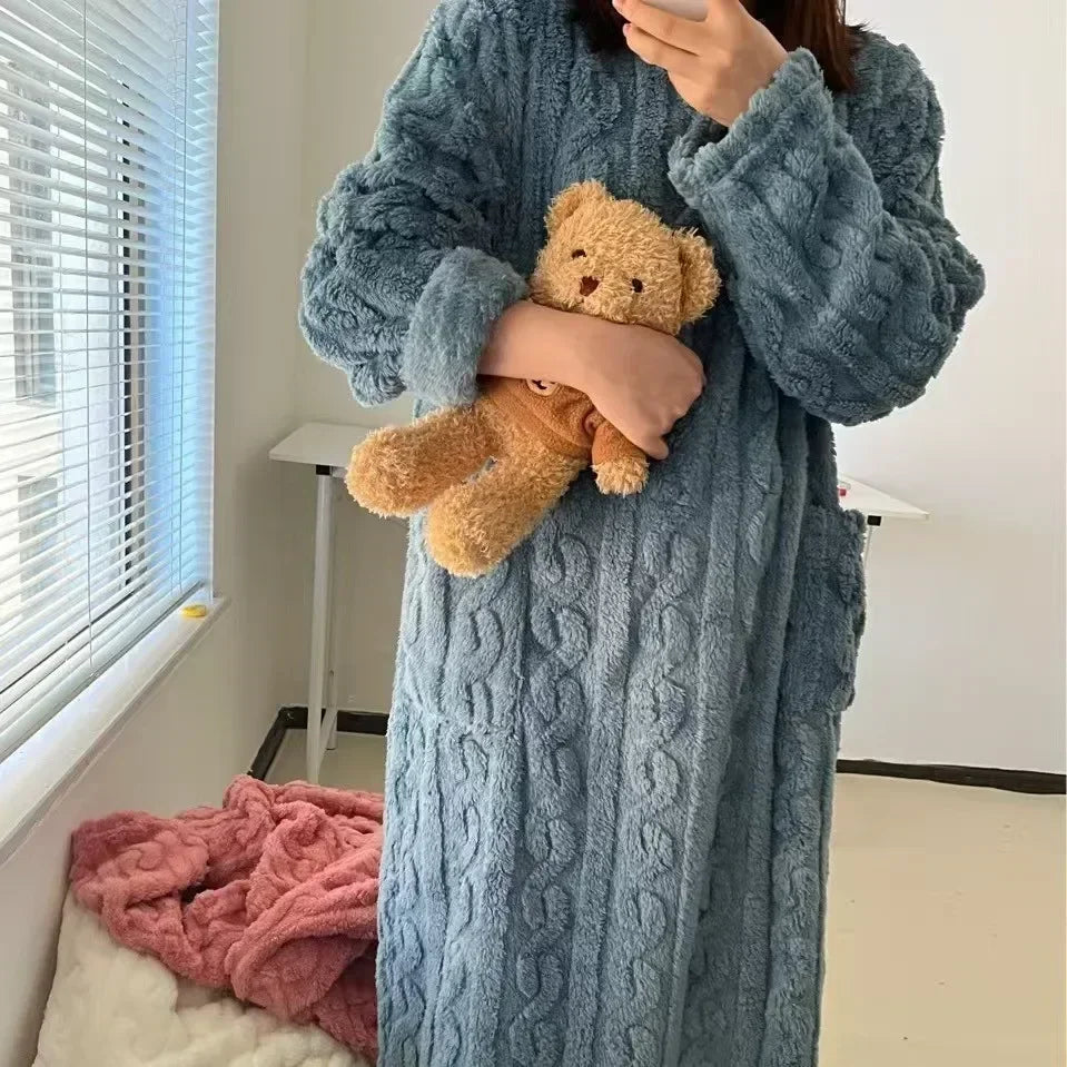 Person wearing a blue cable-knit robe holding a teddy bear in a room with a window and pink blanket.