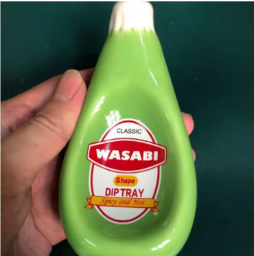 A hand holding a green teardrop-shaped Wasabi dip tray with a white top and a red and white label on a dark green background.