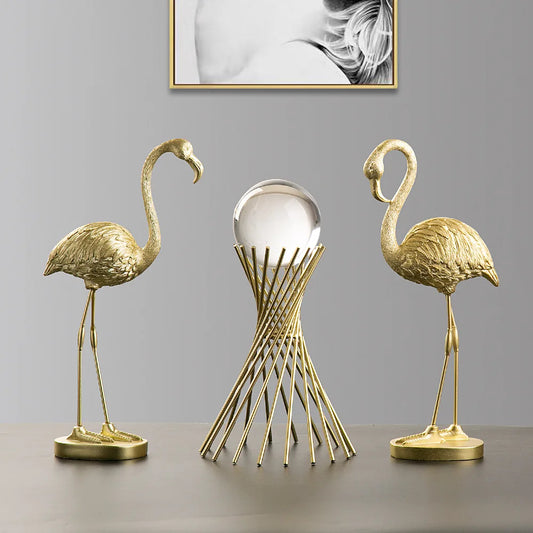 Two gold flamingo sculptures and a central abstract sculpture on a gray surface with a framed picture in the background.