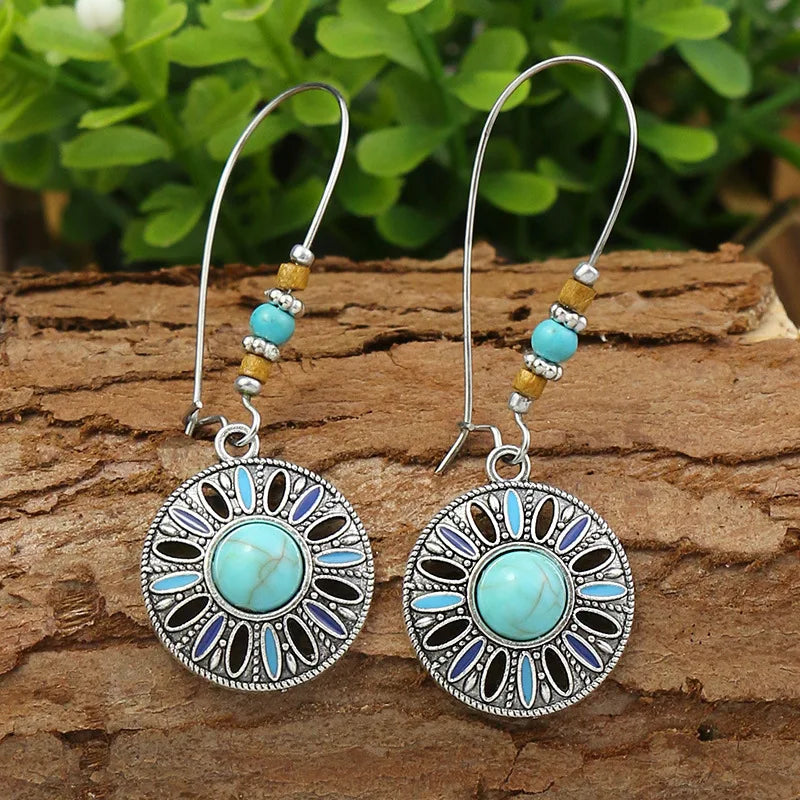 Pair of earrings with turquoise stones on a wooden surface with green leaves in the background