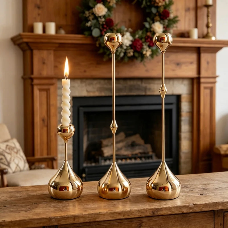Three gold candle holders on a wooden table with a fireplace and Christmas wreath in the background.