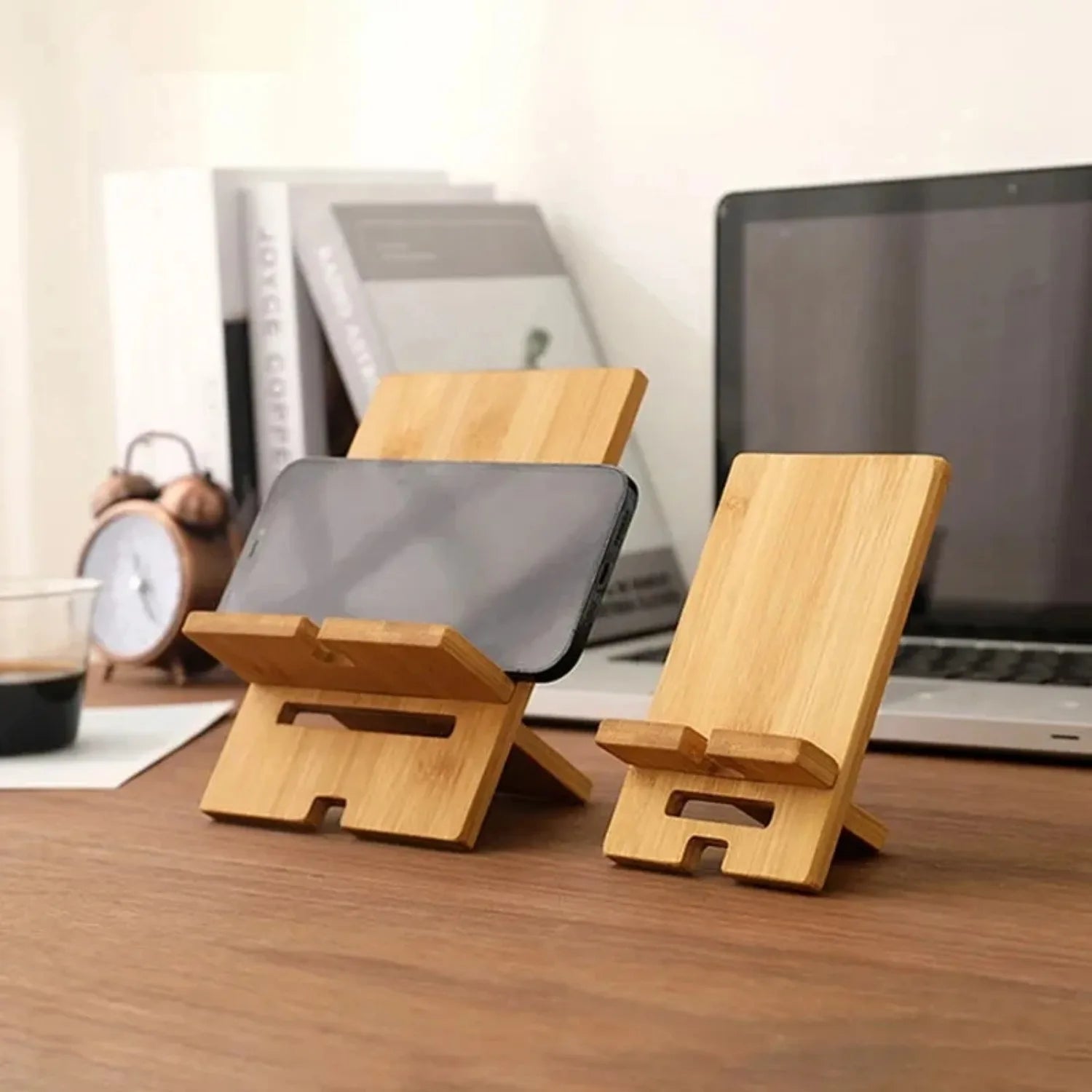 Wooden phone and tablet stands on a desk with a laptop and alarm clock in the background.