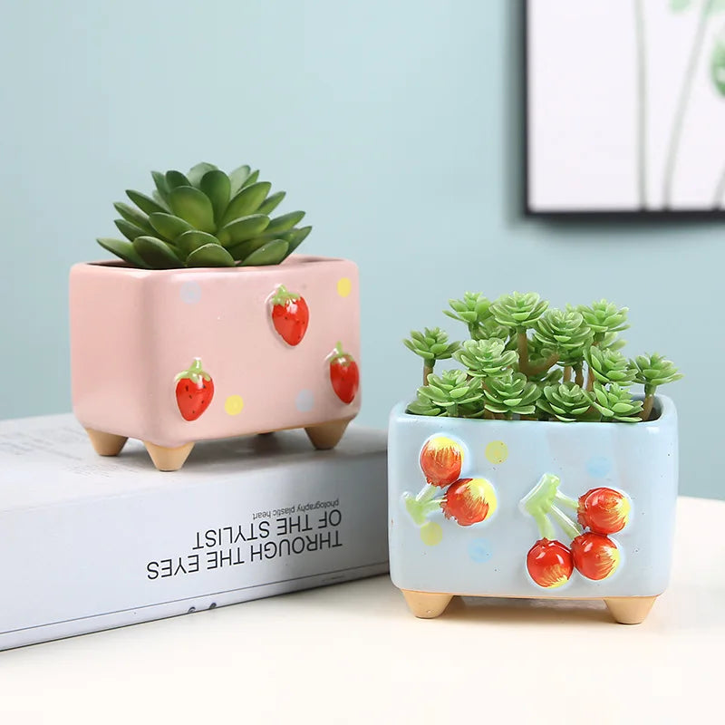 Two small potted plants in decorative pots with strawberry design on a light blue background.