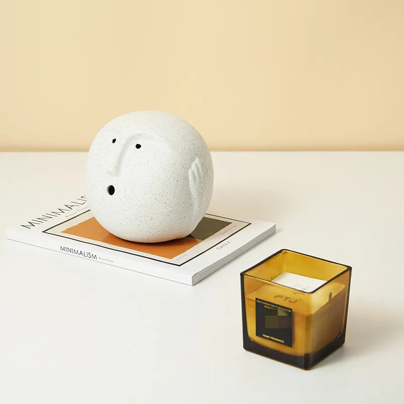 Abstract white ceramic face ornament styled displayed on a book on a whote table next to a candle