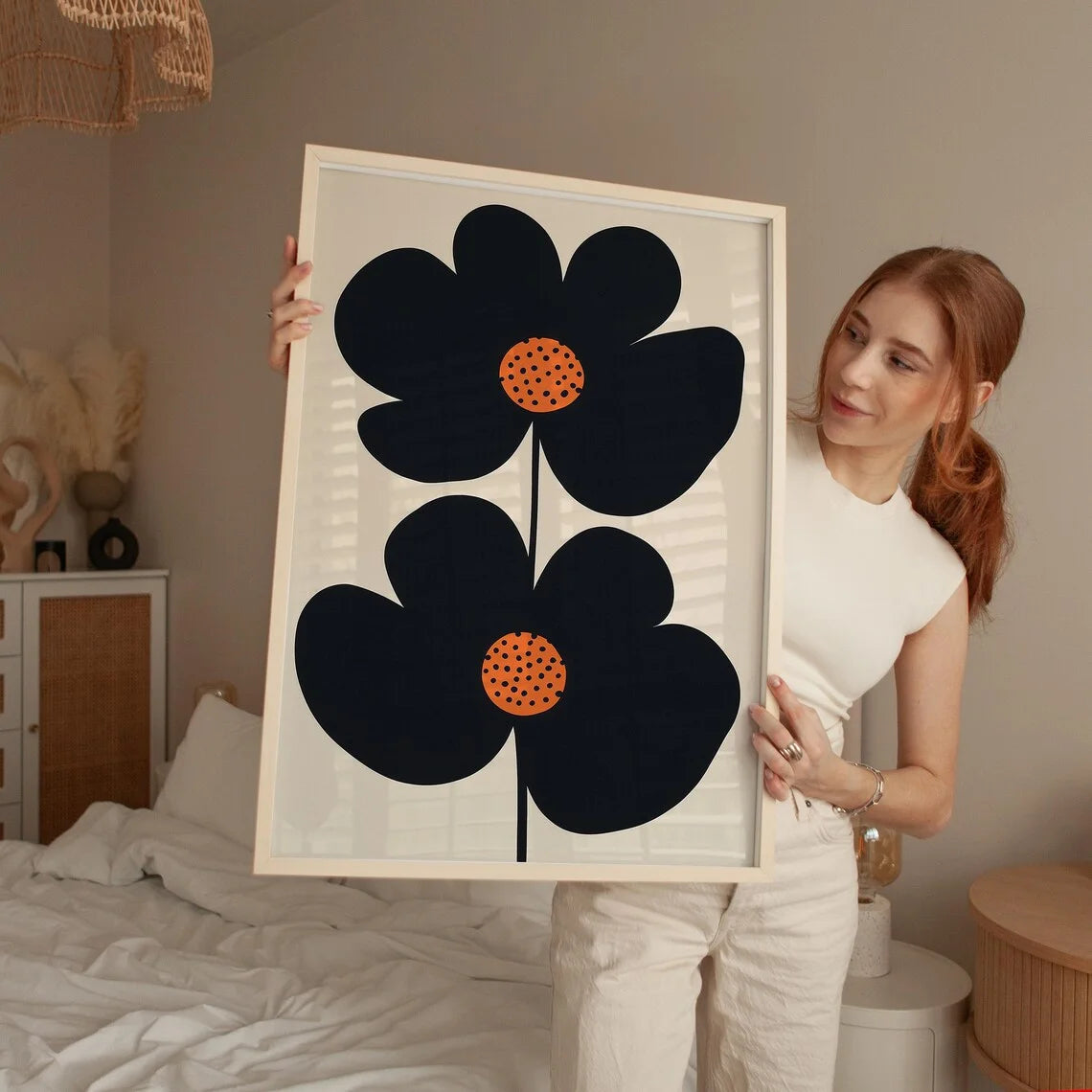 Woman holding a framed artwork of black flowers with orange centers in a bedroom setting.