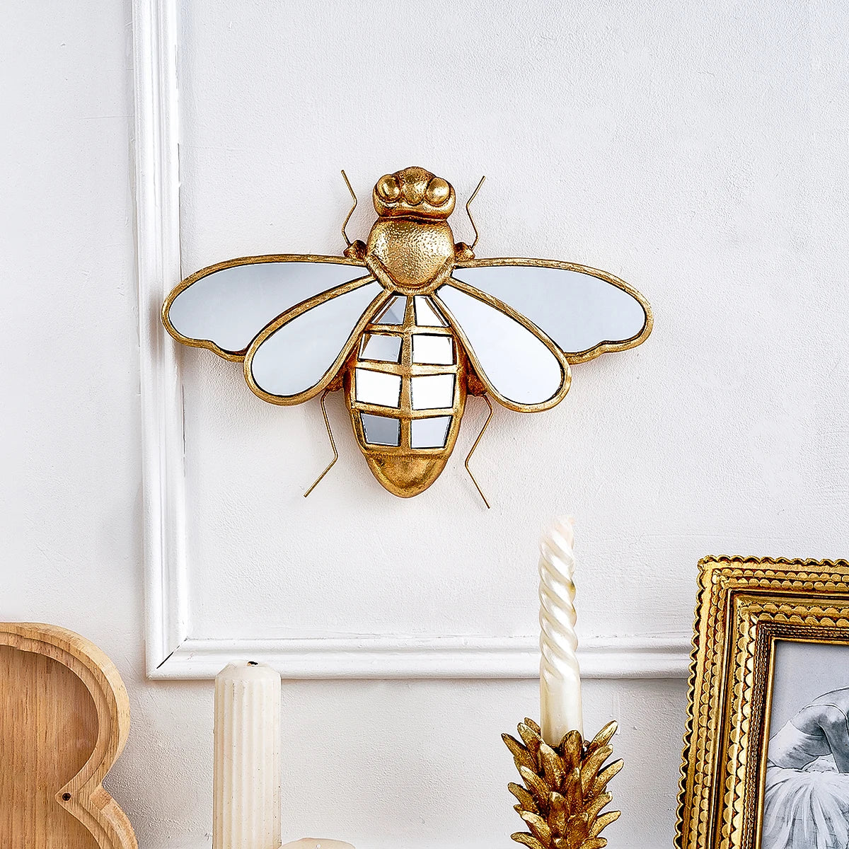 Decorative gold bee wall mirror on a white wall with candles and a photo frame.