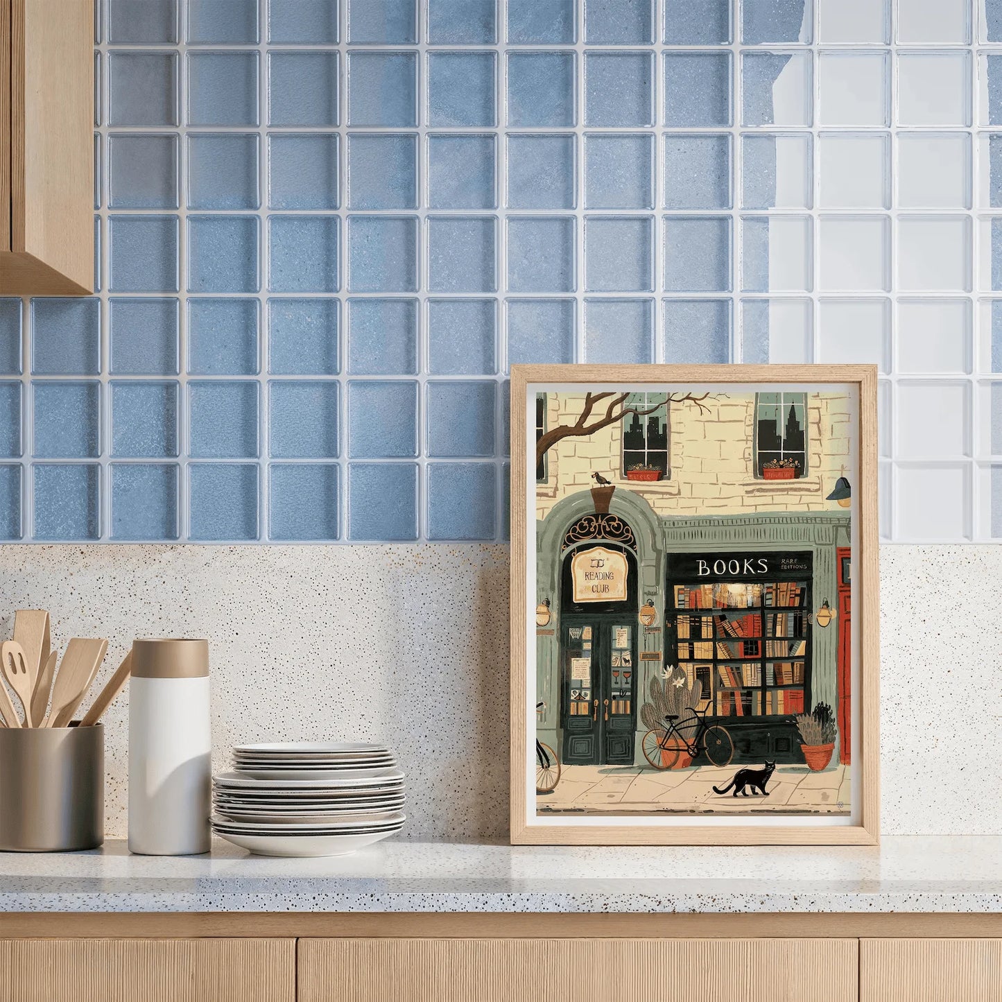 Framed artwork of a bookshop scene on a kitchen counter with blue tiled wall in the background