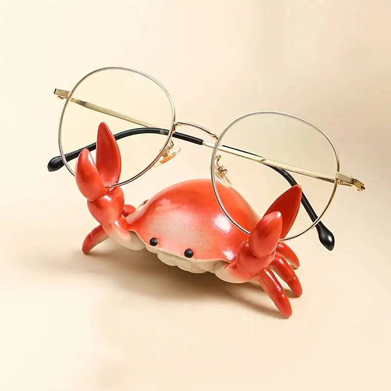 Cute crab-shaped desktop stand holding a smartphone, glasses and stationery, available in colourful finishes for home and office organisation.
