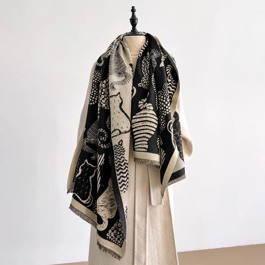 Oversized black and tan cat print shawl scarf with illustrated feline patterns and fringed edges, draped over a neutral coat on a mannequin.