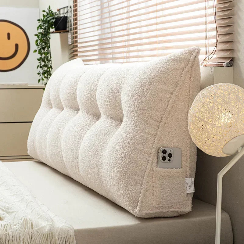 Beige wedge pillow on a bed with a lamp and window blinds in the background
