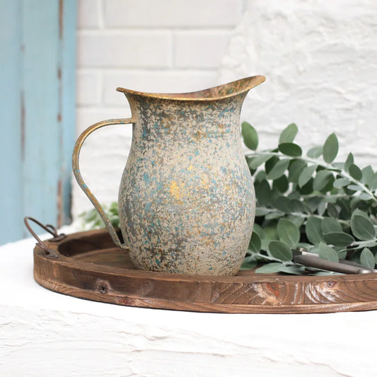 Vintage-style metal pitcher on a wooden tray with greenery in the background