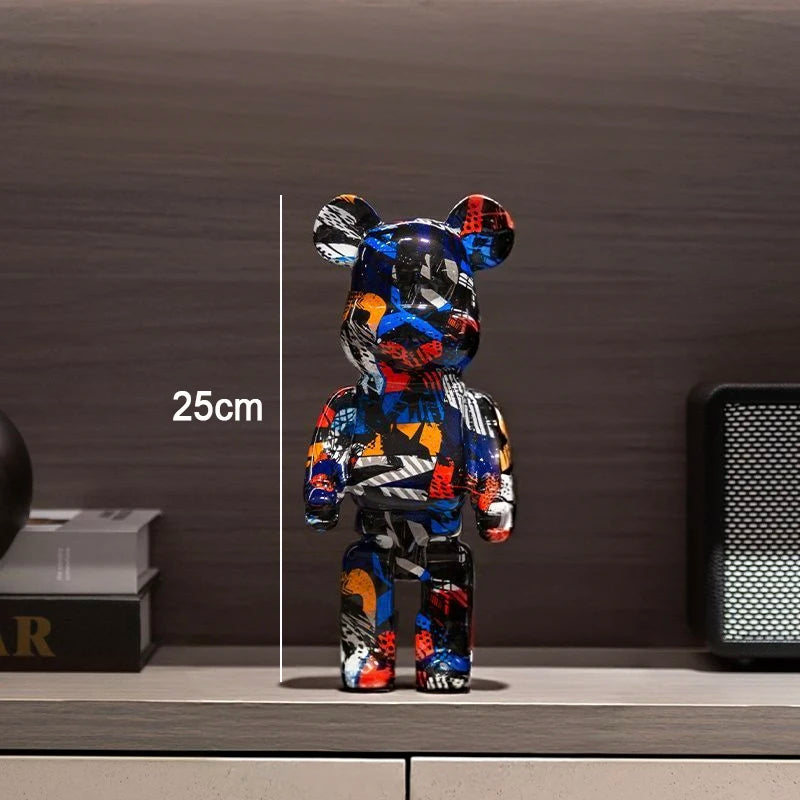 A multicolor graffiti-patterned bear figure stands on a shelf in an indoor setting, with a vertical measurement line and the text '25cm' indicating height. The background includes a dark wood wall, books, and a speaker.
