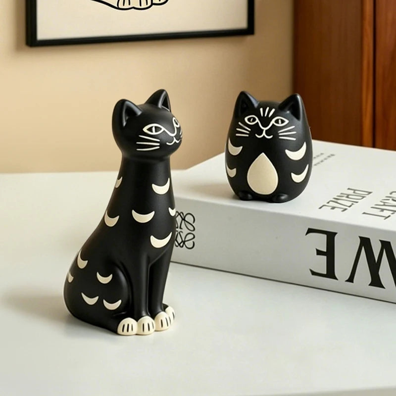 Chubby and tall sitting black cat figurines with cream crescent moon patterns displayed together on a wooden cabinet.
