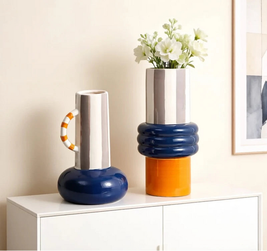 Two ceramic vases on a white cabinet: a handled vase with a rounded blue base and grey striped neck, and a stacked vase with blue rings, an orange base and white flowers.