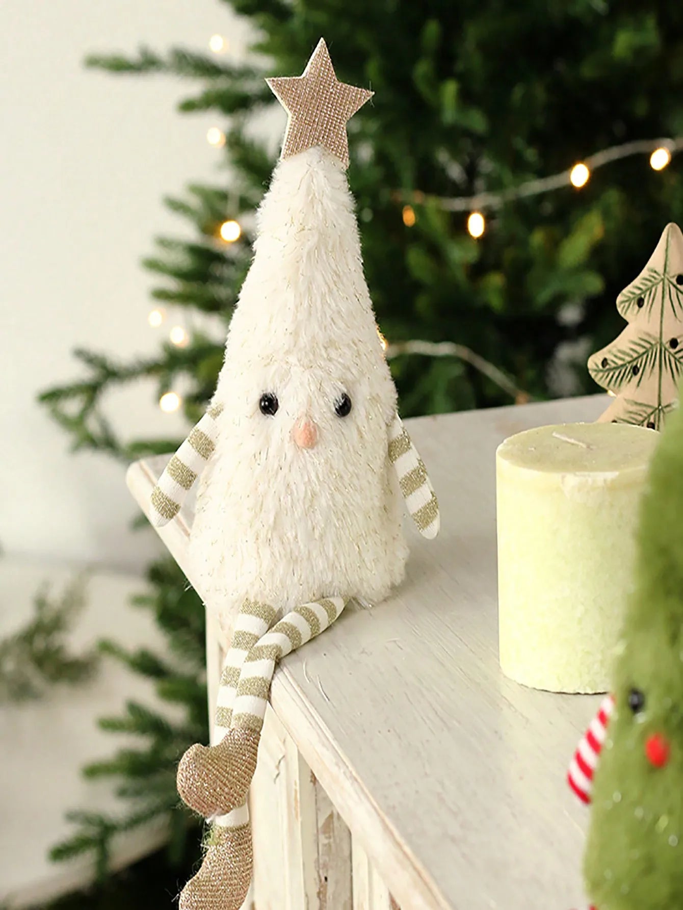 White plush Christmas tree with striped sleeves and a star on top, sitting on a surface with a blurred Christmas tree and candle in the background.