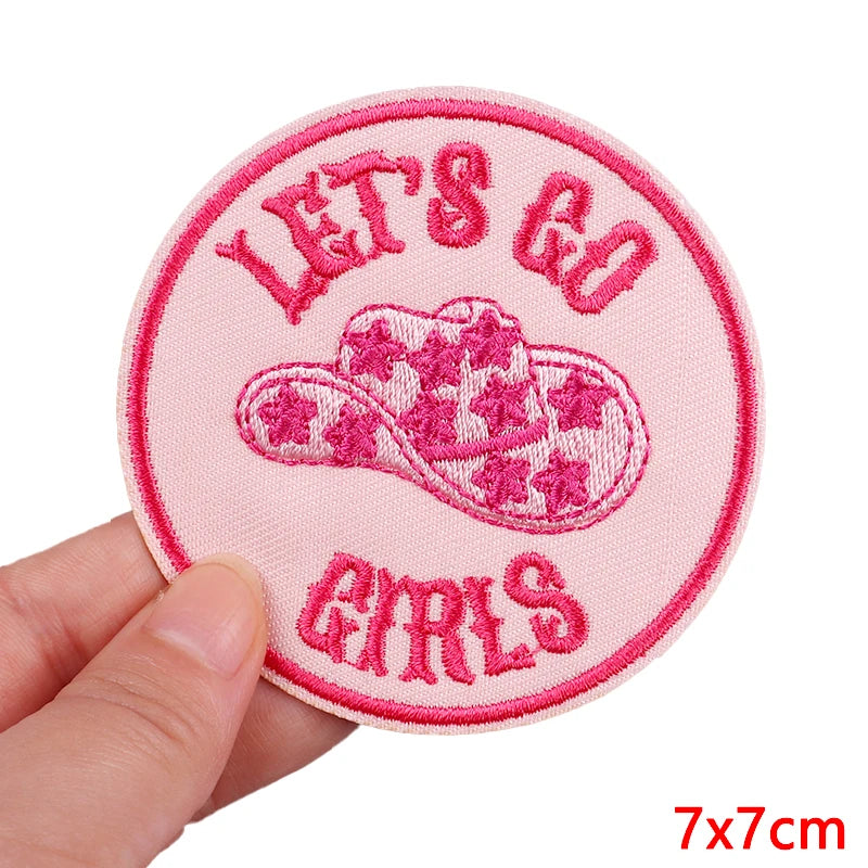 Pink patch with 'Let's Go Girls' text and a cowboy hat design, held by a hand on a white background.