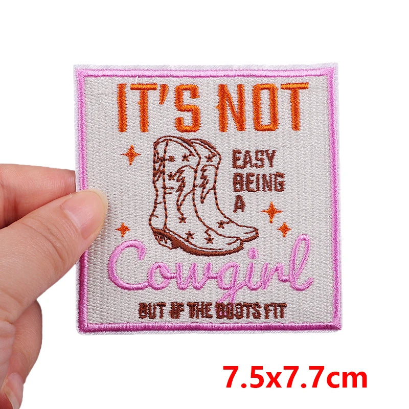 Embroidered patch with cowboy boots and text held by a hand, on a white background.