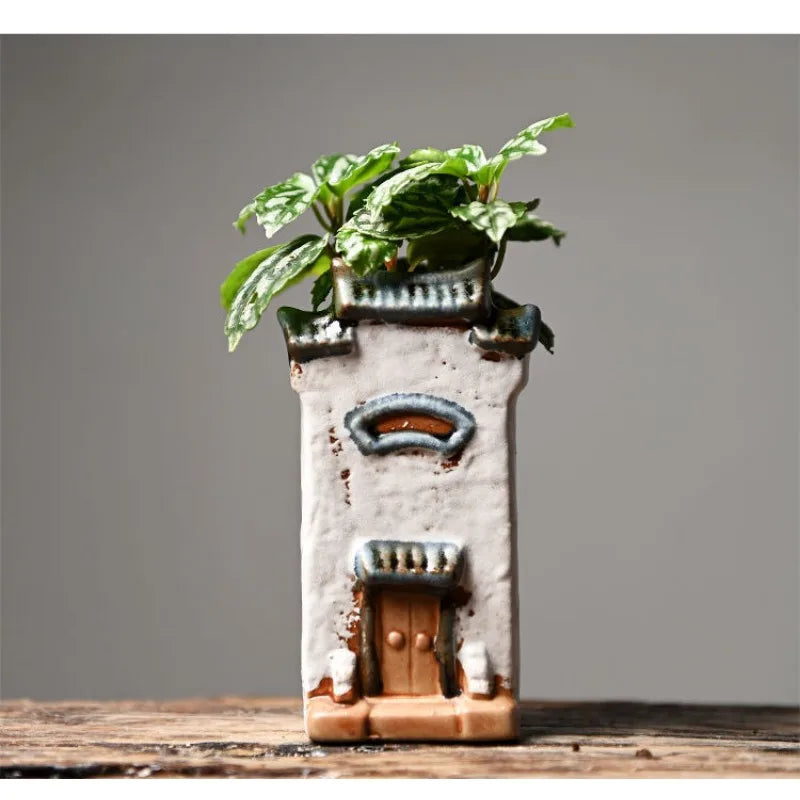 Decorative ceramic plant pot shaped like a small house with a plant on top against a gray background.