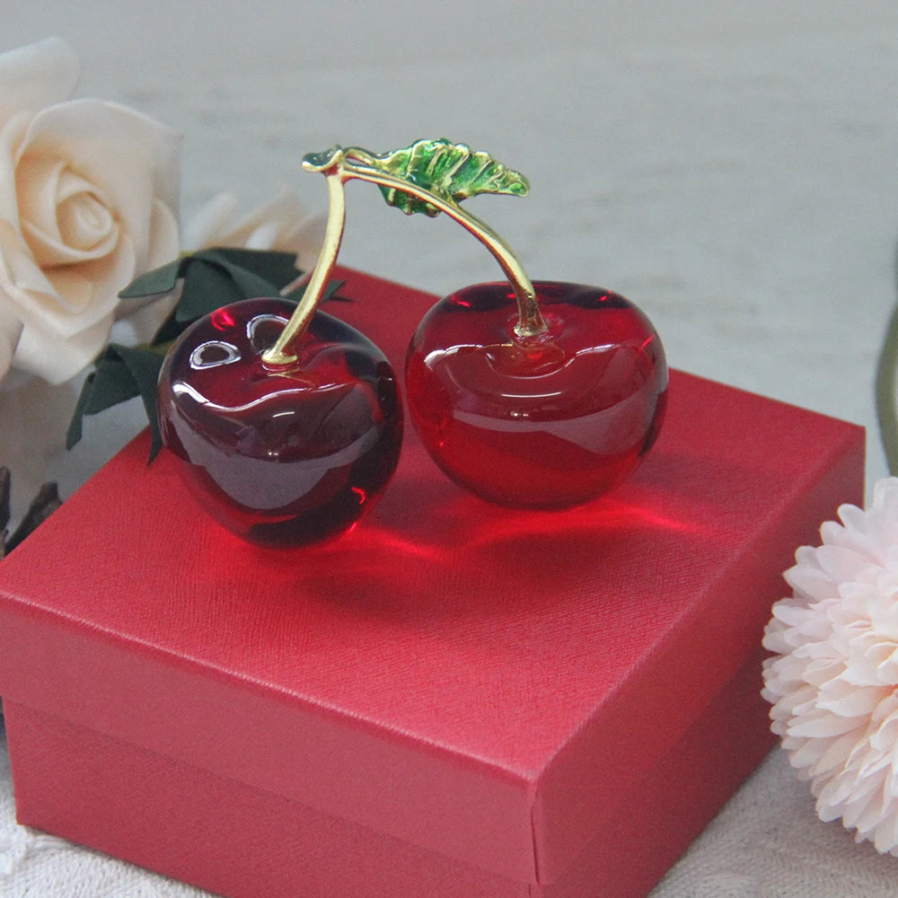 Two red glass cherries with gold stems on a red box, surrounded by flowers.