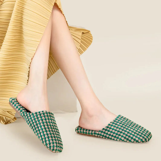 A person wearing green and beige woven mule slippers with a pleated yellow skirt, shown from the knees down against a light beige studio background.