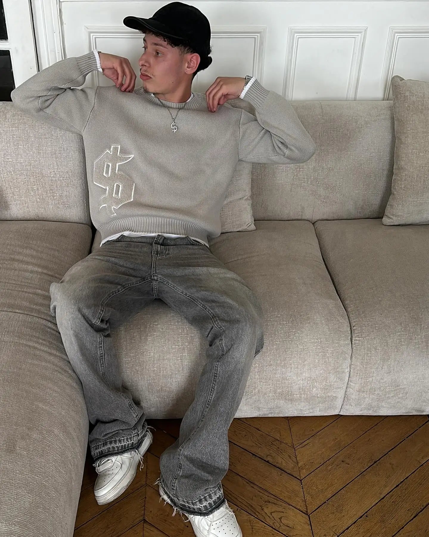 Person sitting on a beige sofa wearing a gray sweater and jeans in a room with wooden flooring.
