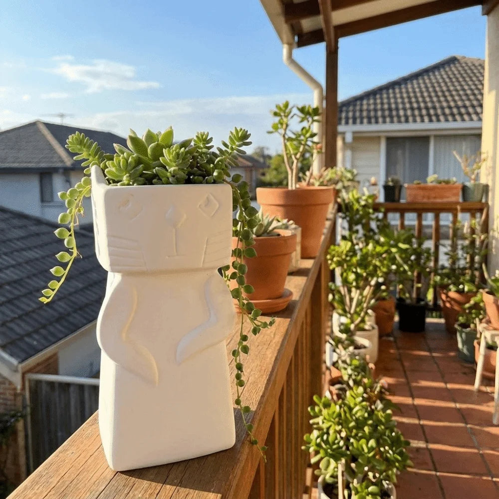 A white ceramic planter shaped like a cat sitting on a wooden balcony railing, filled with green succulent plants. The balcony is surrounded by other potted plants and residential houses under a blue sky.