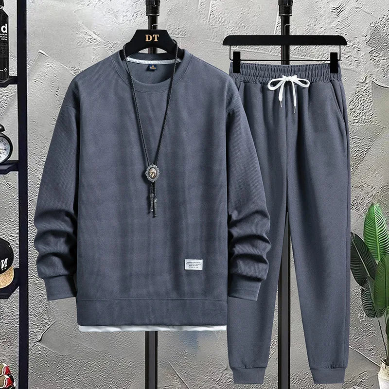 Gray tracksuit with a necklace on a hanger against a textured wall.