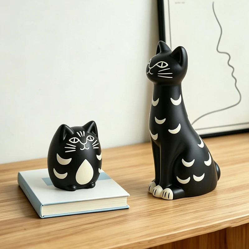 Two black cat figurines with white crescent moon patterns, one larger seated figure on a wooden shelf and one smaller standing figure resting on a book, against a light wall with framed wall art.