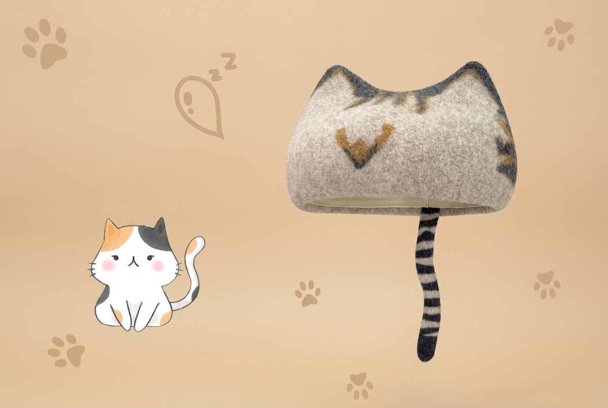 A wool cat-shaped hat with a striped tail, shown on a beige background with paw prints and a small cat illustration. 