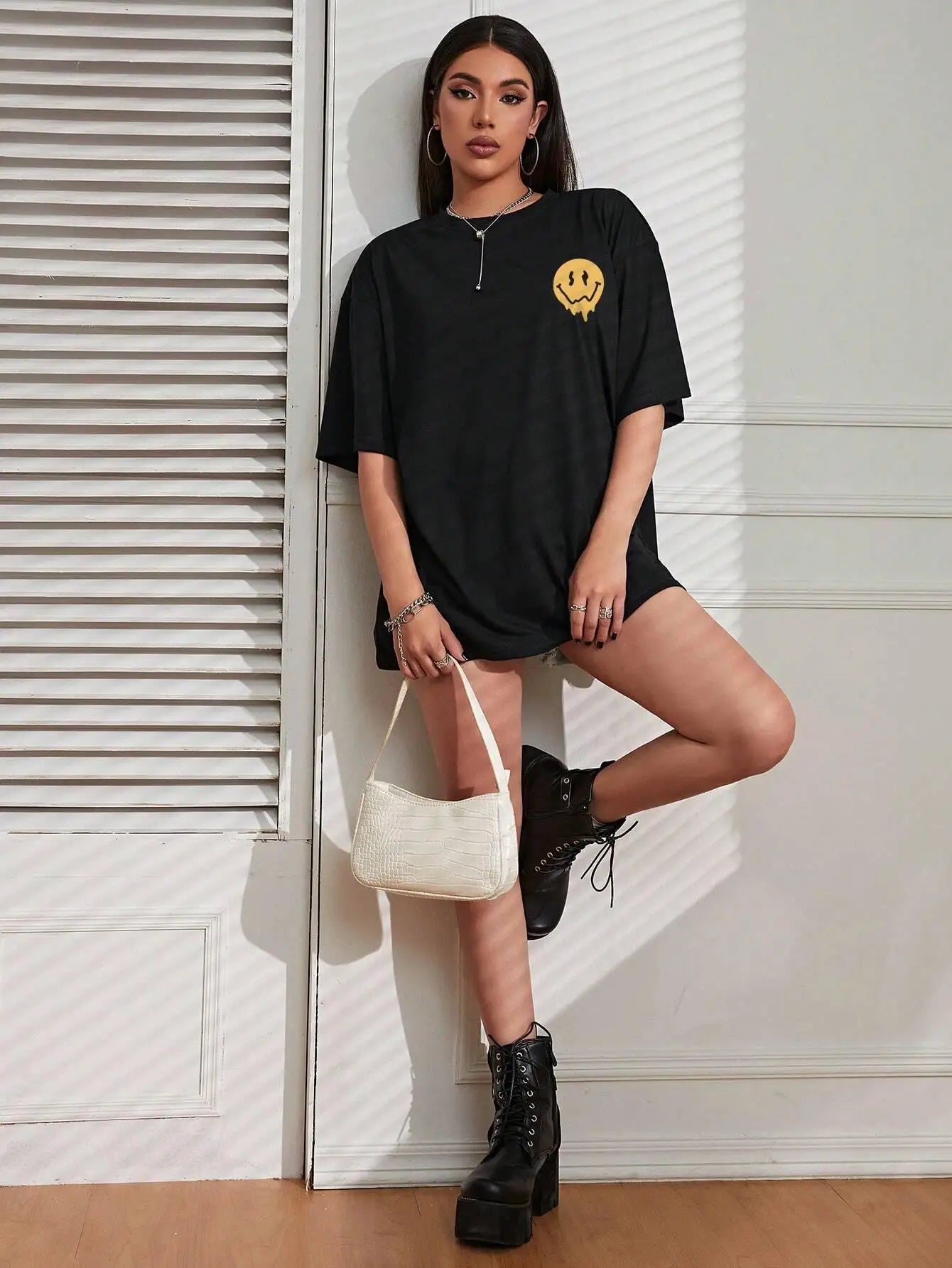 Woman wearing a black oversized t-shirt with a yellow logo, holding a white handbag against a white wall.