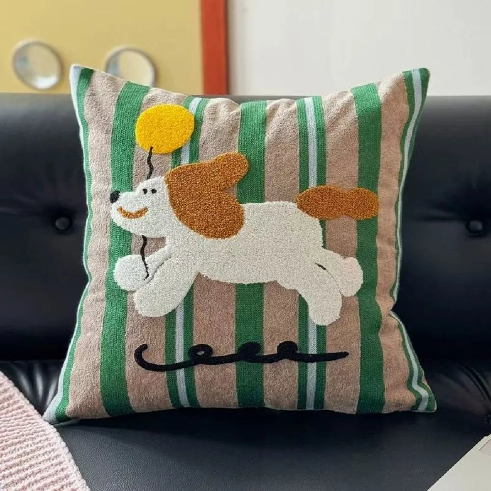 A square throw pillow with a green and beige striped pattern, featuring a raised, textured applique of a white dog with brown ears and tail, holding a yellow balloon. The pillow is resting on a black leather sofa.