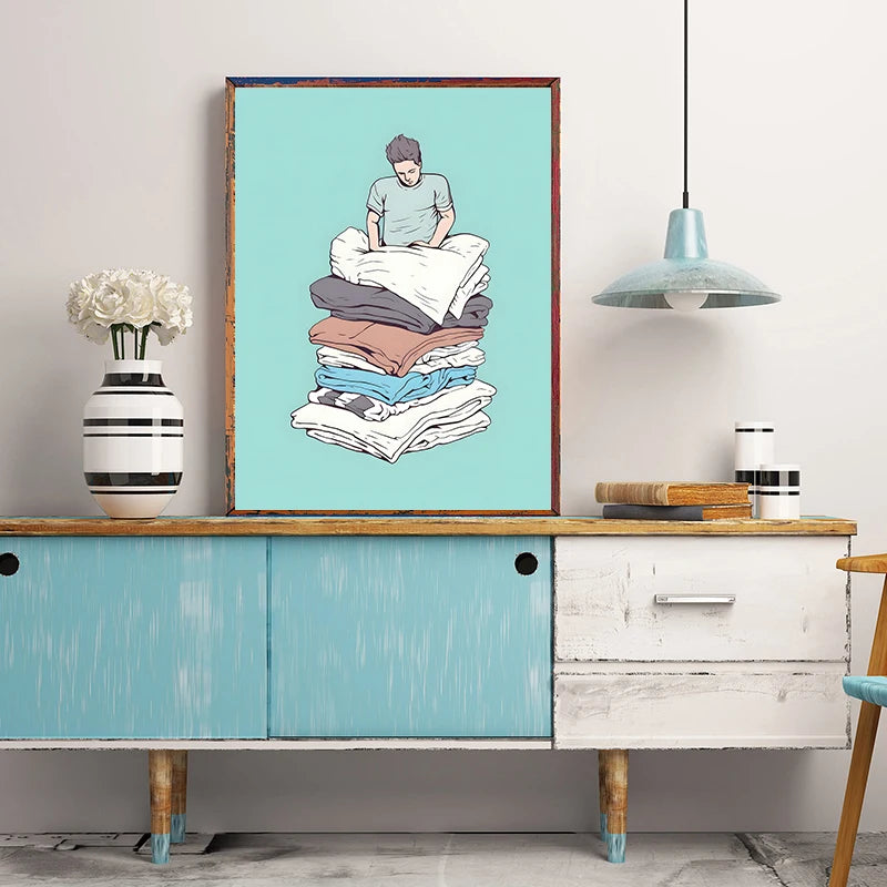 Framed artwork of a person sitting on folded clothes above a wooden cabinet with a lamp and vase.