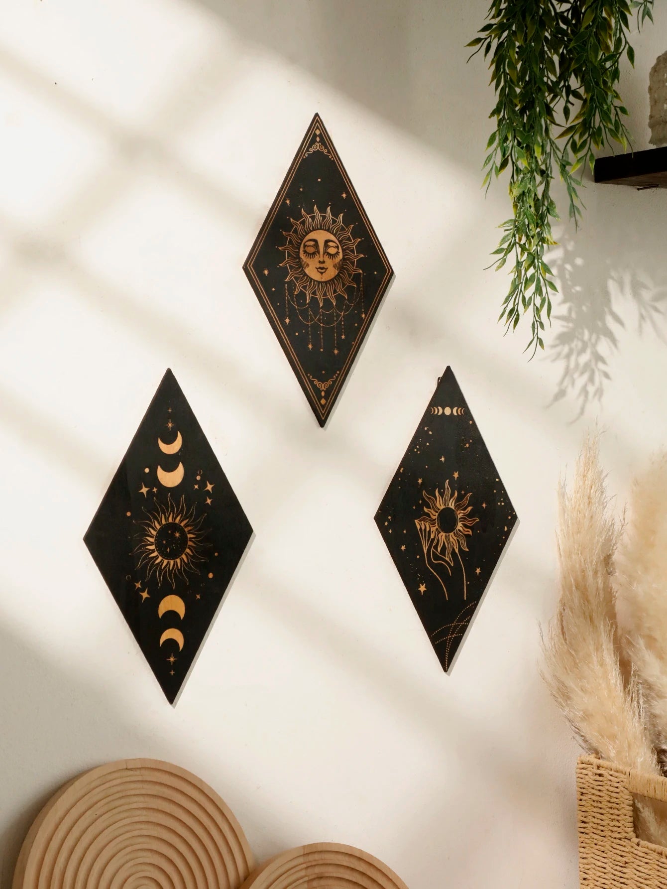 Three black diamond-shaped wall art pieces with gold celestial designs are displayed on a white wall in a styled room setting with soft window shadows, a hanging green plant, and dried pampas grass.