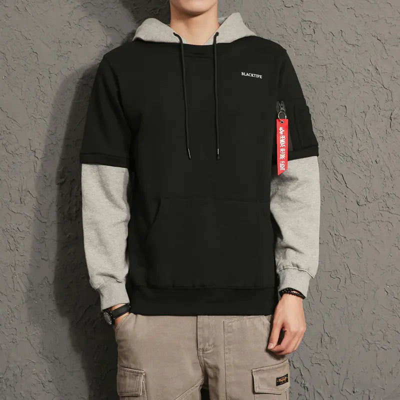 Person wearing a black and gray hoodie with a red tag against a gray textured wall