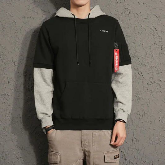 Person wearing a black and gray hoodie with a red tag against a gray textured wall