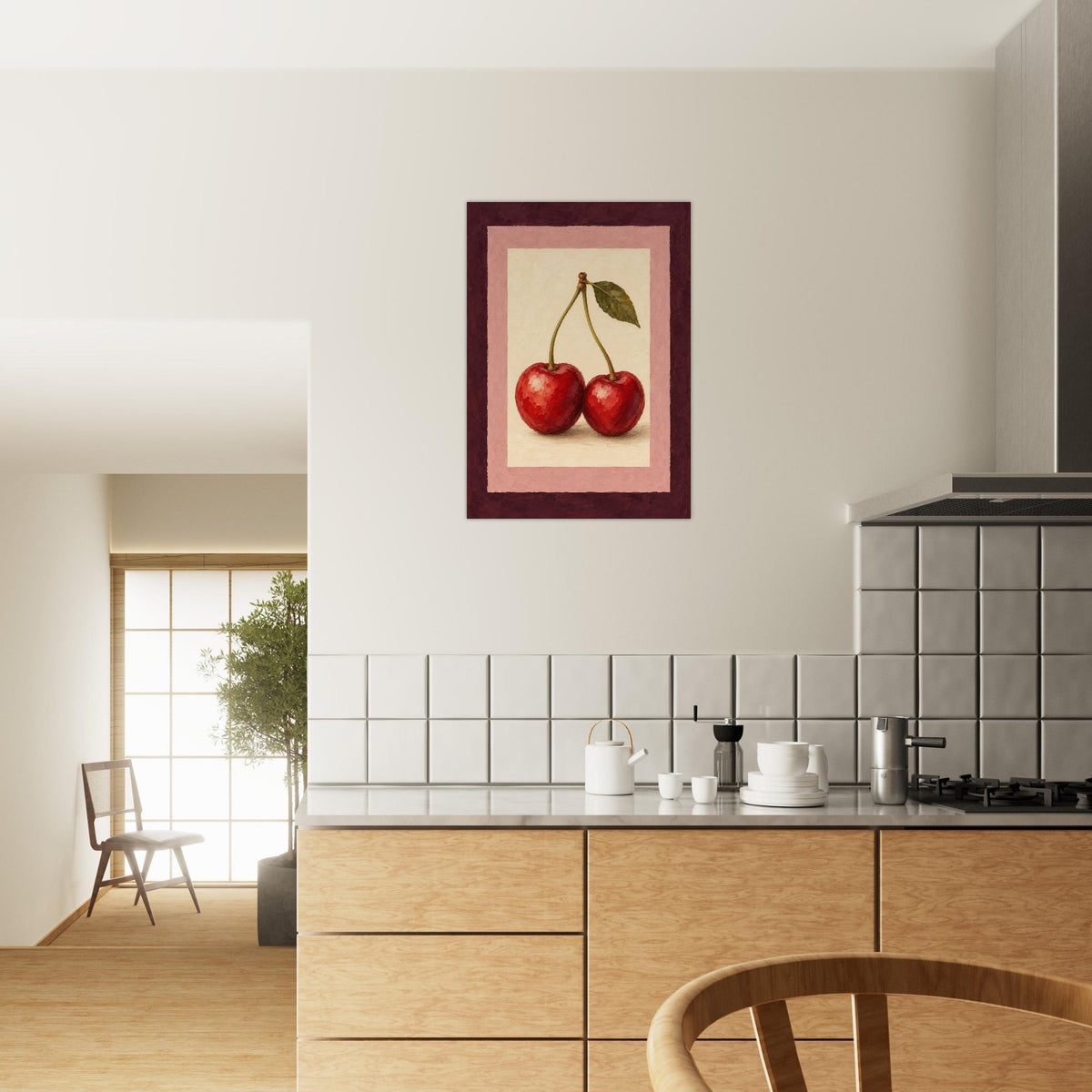 A framed wall art print depicting two red cherries with green leaves, hung on a beige wall above a modern kitchen with light wood cabinets and white tiles.