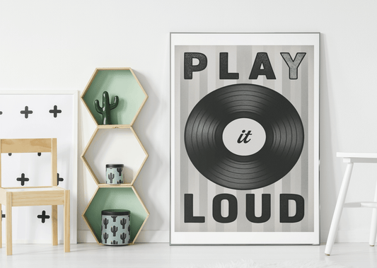 Framed black and white vinyl record art print with 'Play It Loud' typography displayed in a modern minimalist kids room with hexagonal shelves and cactus decor.