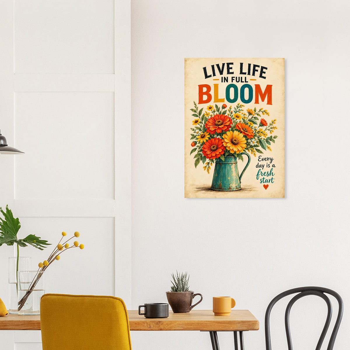 A vertical metal wall art print featuring a colorful bouquet of flowers in a green pitcher with the text 'LIVE LIFE IN FULL BLOOM' and 'Every day is a fresh start', displayed on a white wall above a wooden dining table in a modern room setting.