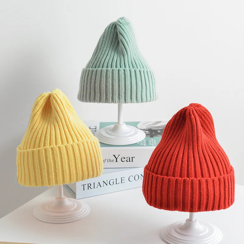 Three knit beanie hats in yellow, green, and red on white stands against a light gray background.