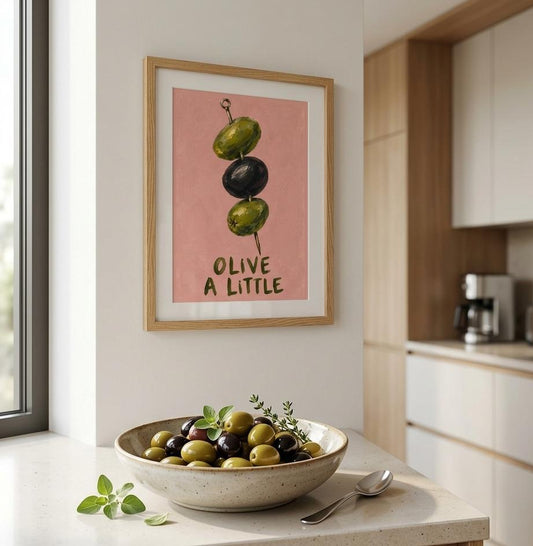 A framed art print featuring three olives on a pink background with the text 'OLIVE A LITTLE' hangs on a kitchen wall above a bowl of fresh olives.